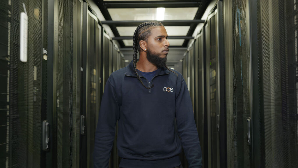 A man with plaited hair and a beard, wearing a dark OCS sweatshirt, stands in a server room aisle surrounded by tall server racks.