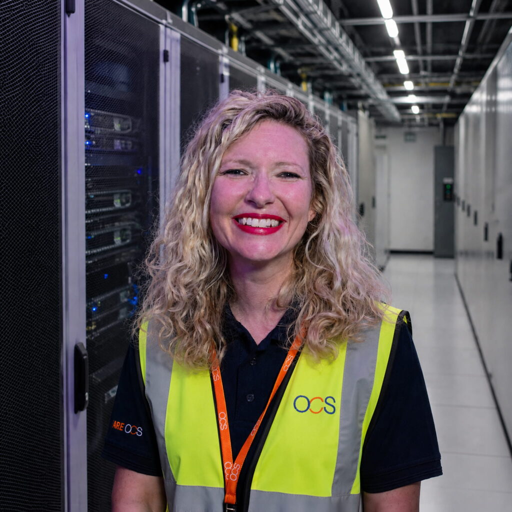 A woman with curly blonde hair, wearing a yellow safety vest and OCS lanyard, smiles whilst standing in a modern server room filled with racks of equipment.
