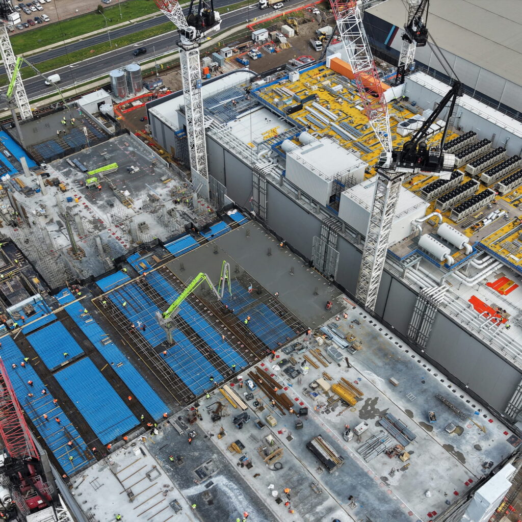 Aerial view of a large construction site with cranes, workers, blue formwork panels, building materials, and partially completed structures. Roads and vehicles are visible in the background.