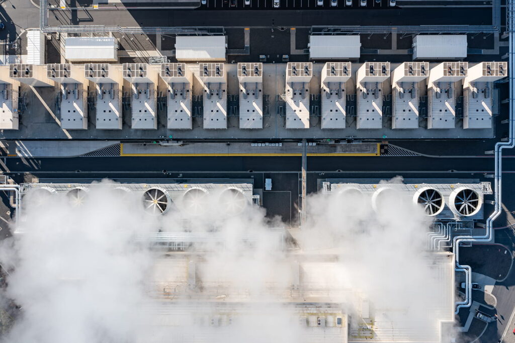 Aerial view of an industrial facility with rows of large rectangular units and cooling fans. Steam or smoke partially obscures the lower section, while pipes and parked cars are visible at the top.