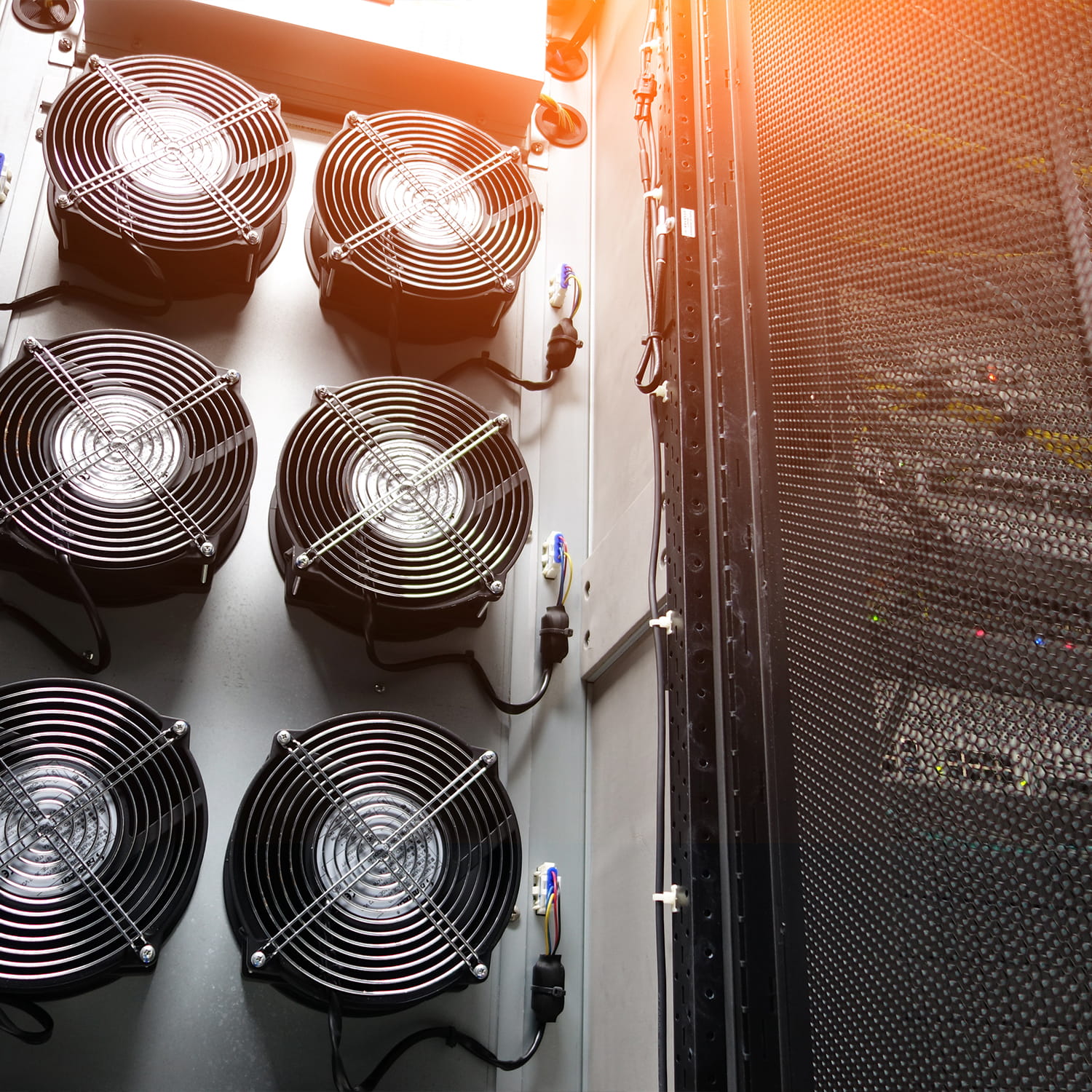 Six large cooling fans mounted vertically in a server room next to a mesh-lined data rack, with bright sunlight streaming in from the upper right corner.
