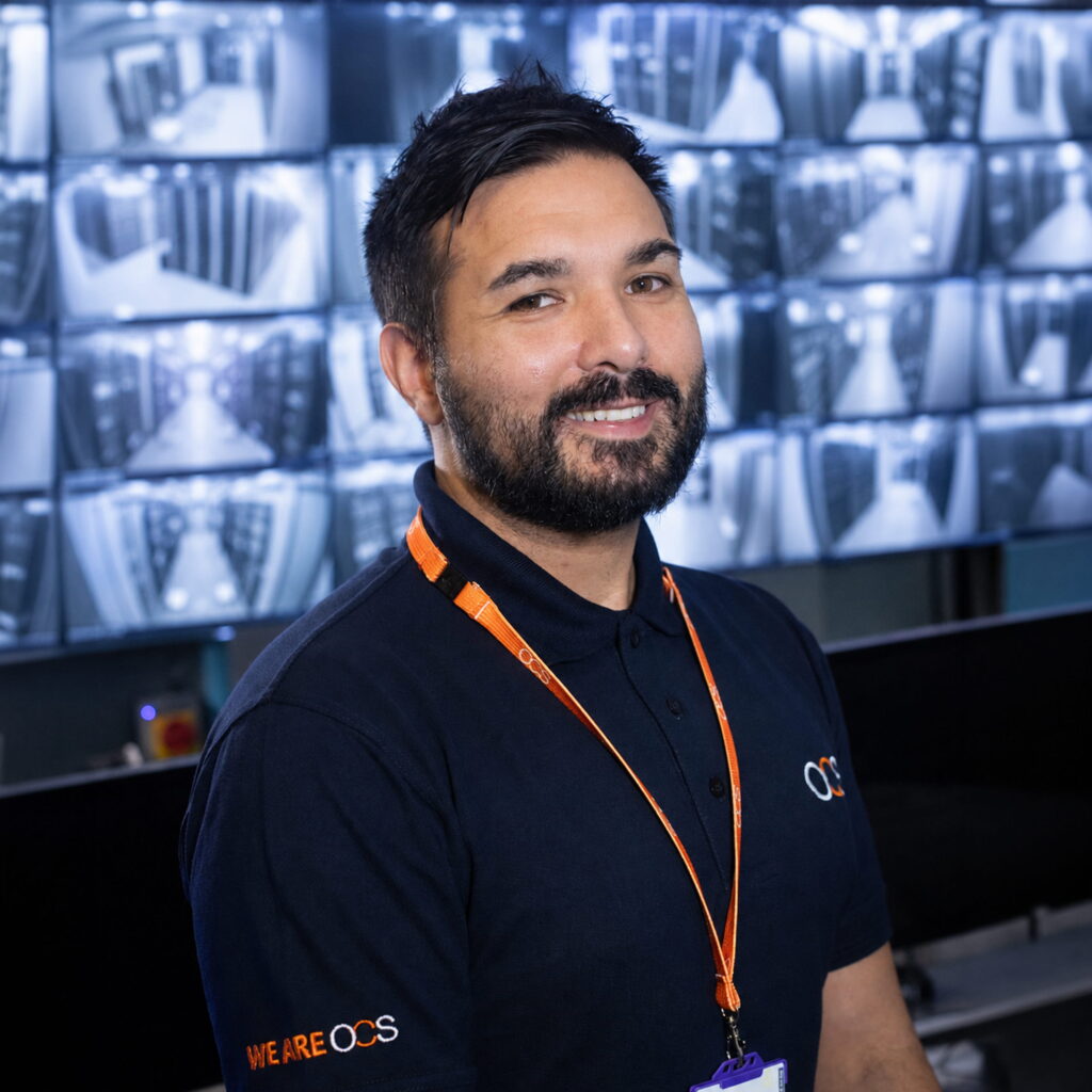A man with dark hair and a beard wears a navy OCS shirt and orange lanyard, smiling in front of multiple security monitors displaying CCTV footage.