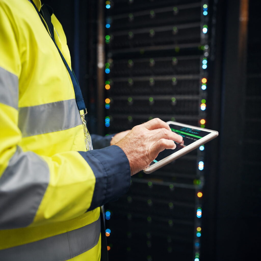 A person in a high-visibility jacket uses a tablet in front of a server rack with glowing indicator lights.