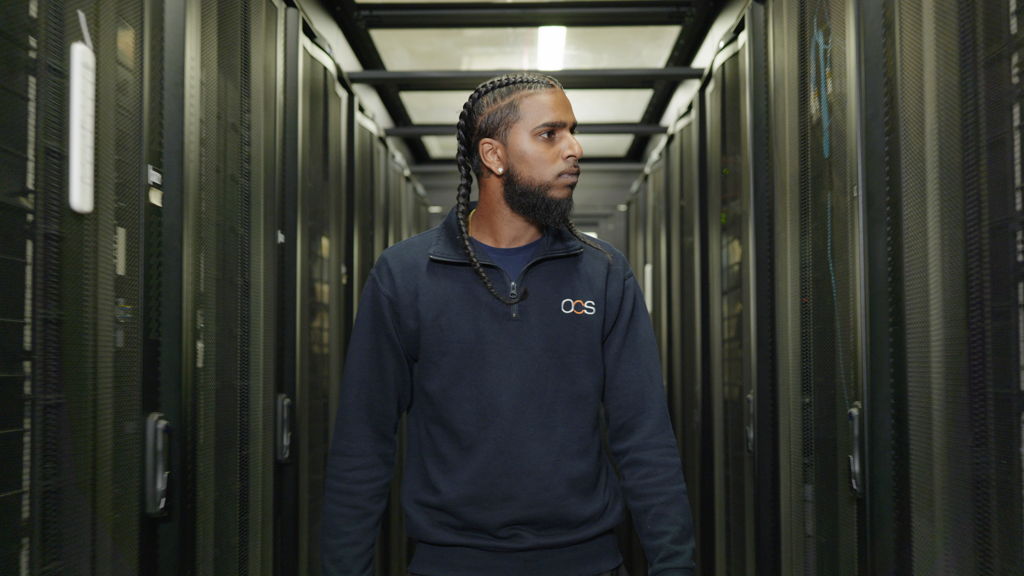 A man with plaited hair and a beard walks through a server room lined with tall black server racks, wearing a dark sweatshirt with an OCS logo on it.