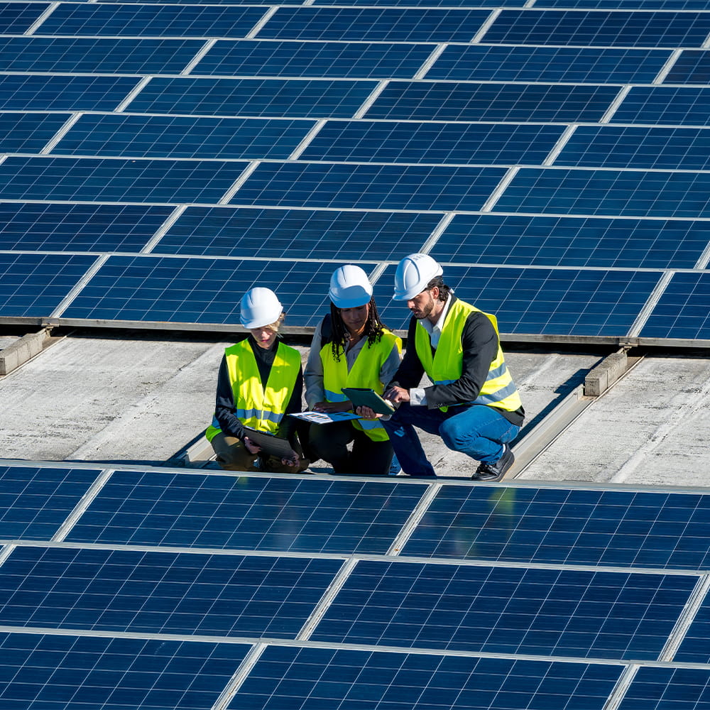 Three people in hard hats and yellow safety vests examine documents while kneeling among rows of solar panels on a rooftop under bright sunlight.