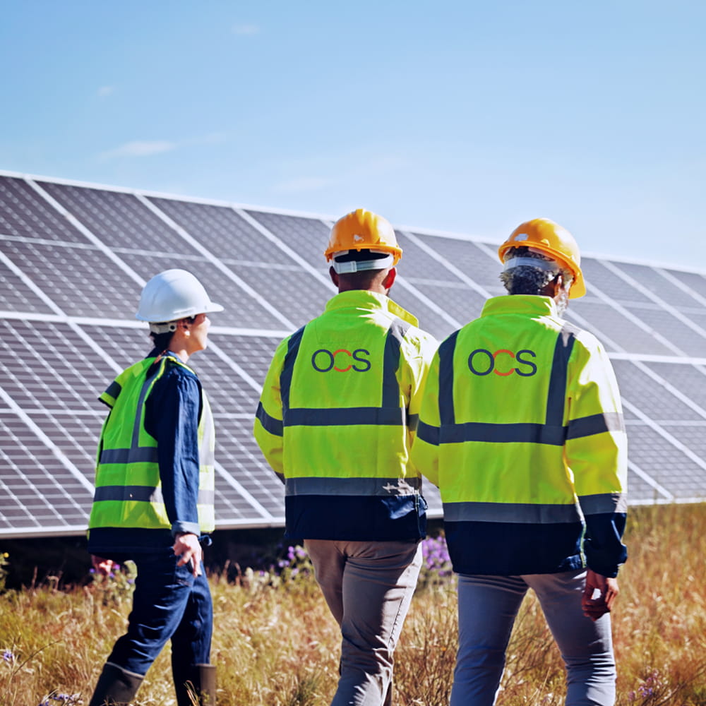 Three workers in safety gear walk beside large solar panels under a clear sky. They wear yellow reflective jackets and hard hats, and OCS is visible on the jackets.