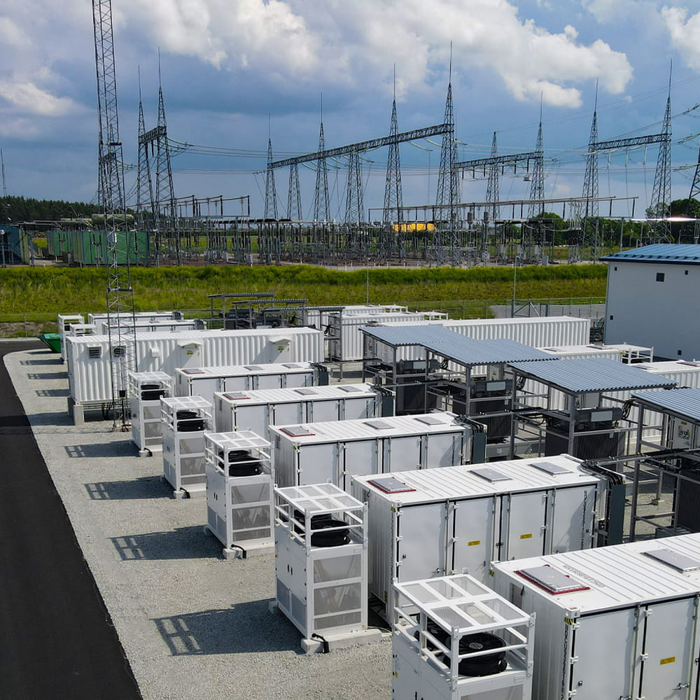 A row of white, container-like battery storage units is set up outdoors near an electrical substation with metal pylons and power lines under a partly cloudy sky.