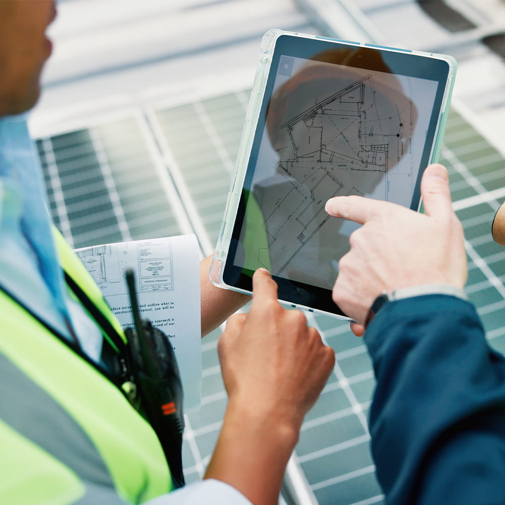Two people in safety vests review architectural plans on a tablet while standing next to solar panels. One person points at the screen, and a paper with diagrams is visible in the other’s hand.
