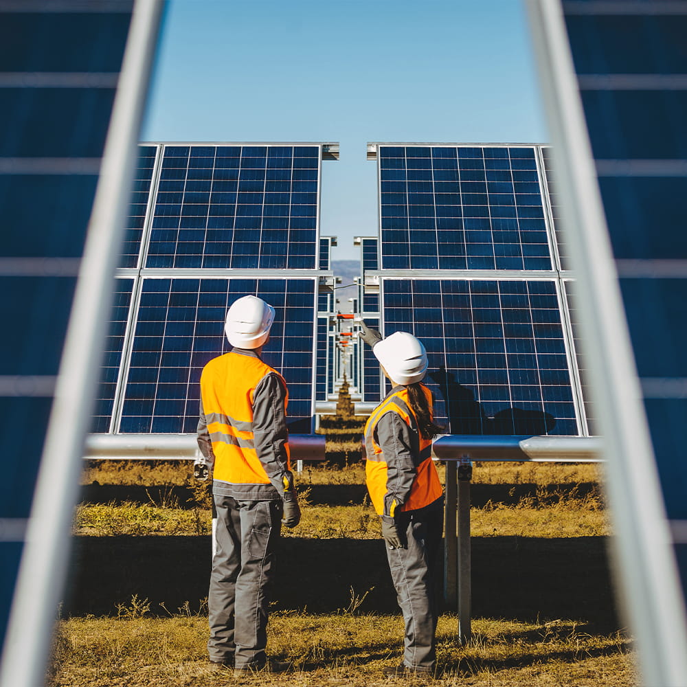 Two workers in orange safety vests and white helmets stand in front of large solar panels outdoors, inspecting or discussing the solar power installation on a sunny day.