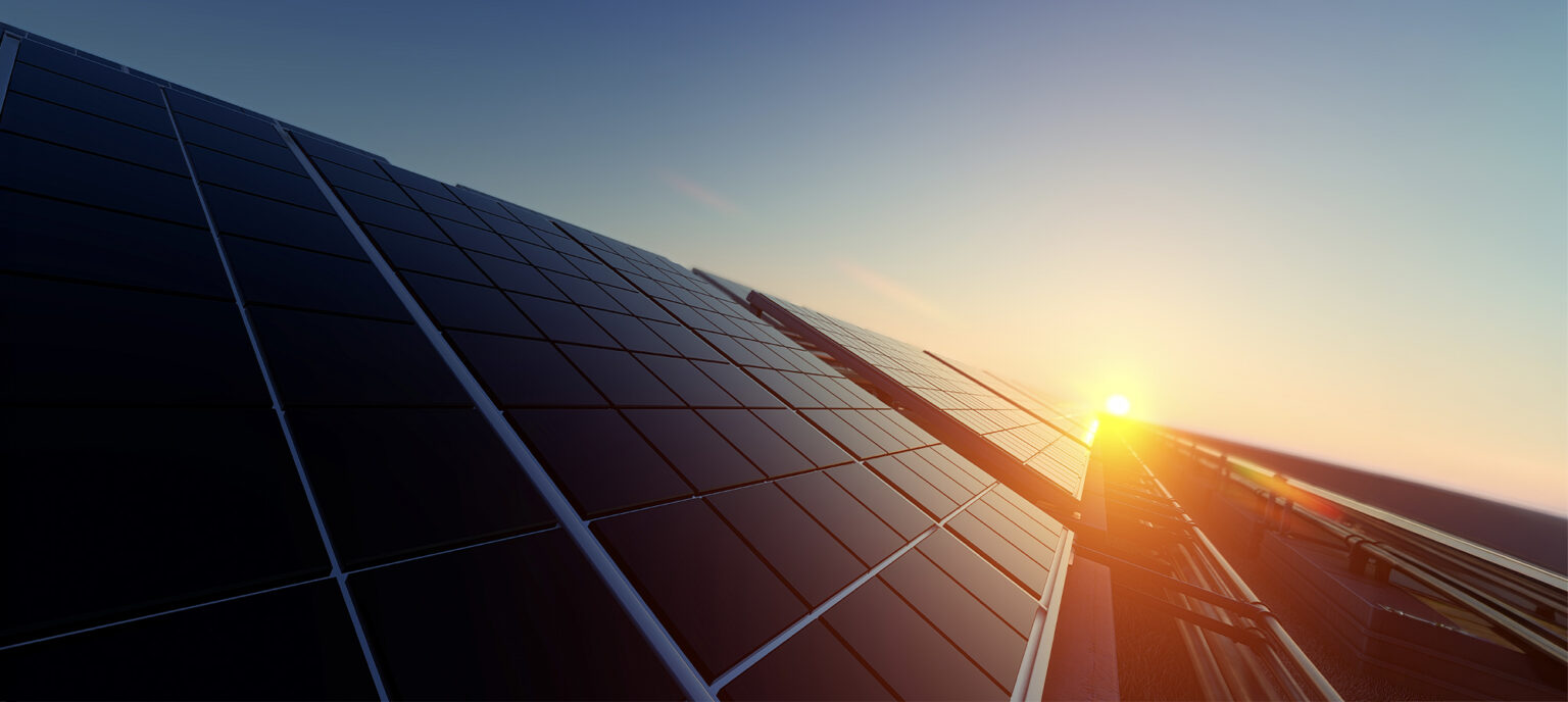 Rows of solar panels are mounted on a roof, reflecting sunlight as the sun sets or rises in the background, creating a bright, warm glow.