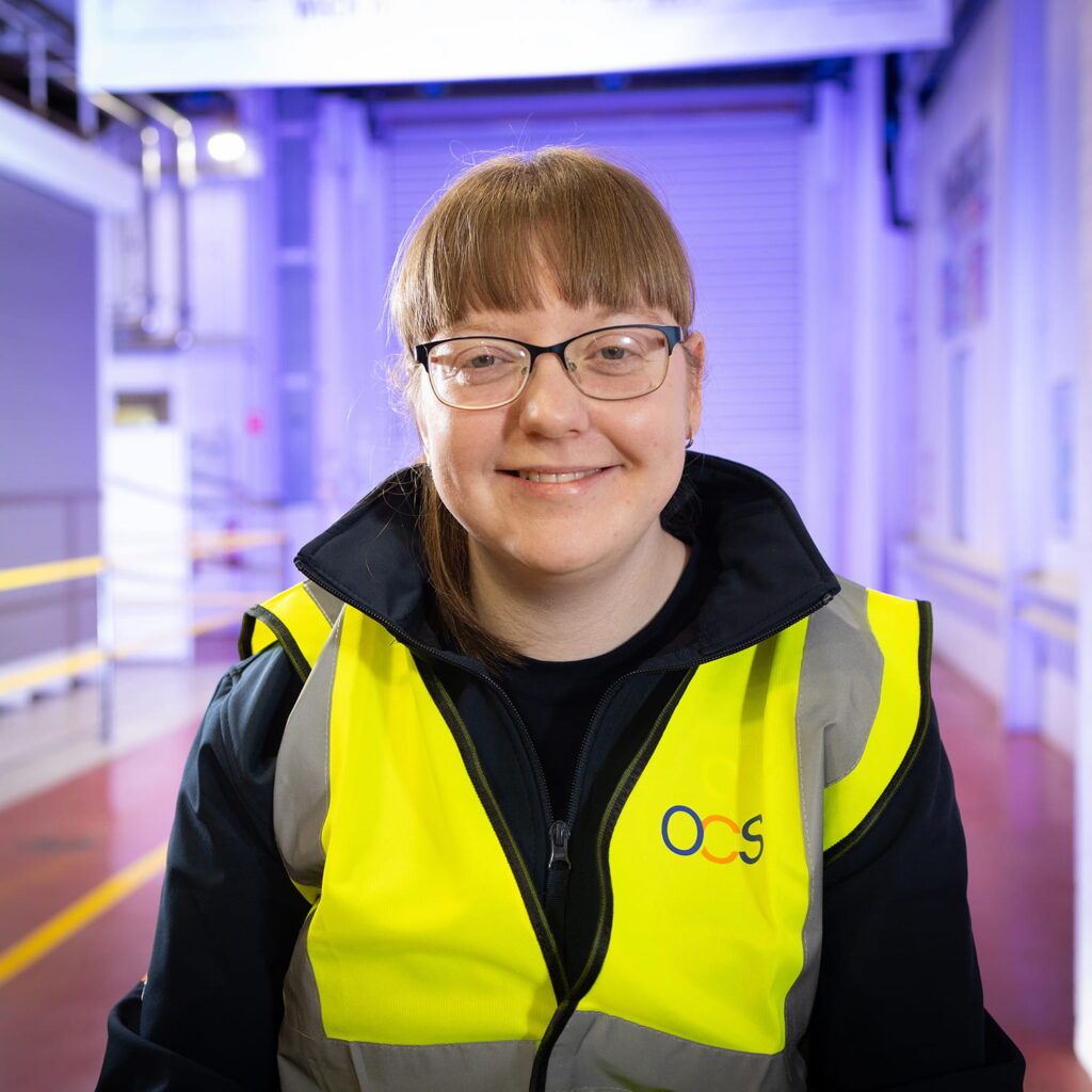 A woman wearing glasses and a high-visibility yellow vest with the OCS logo smiles while standing in a brightly lit industrial or warehouse setting.