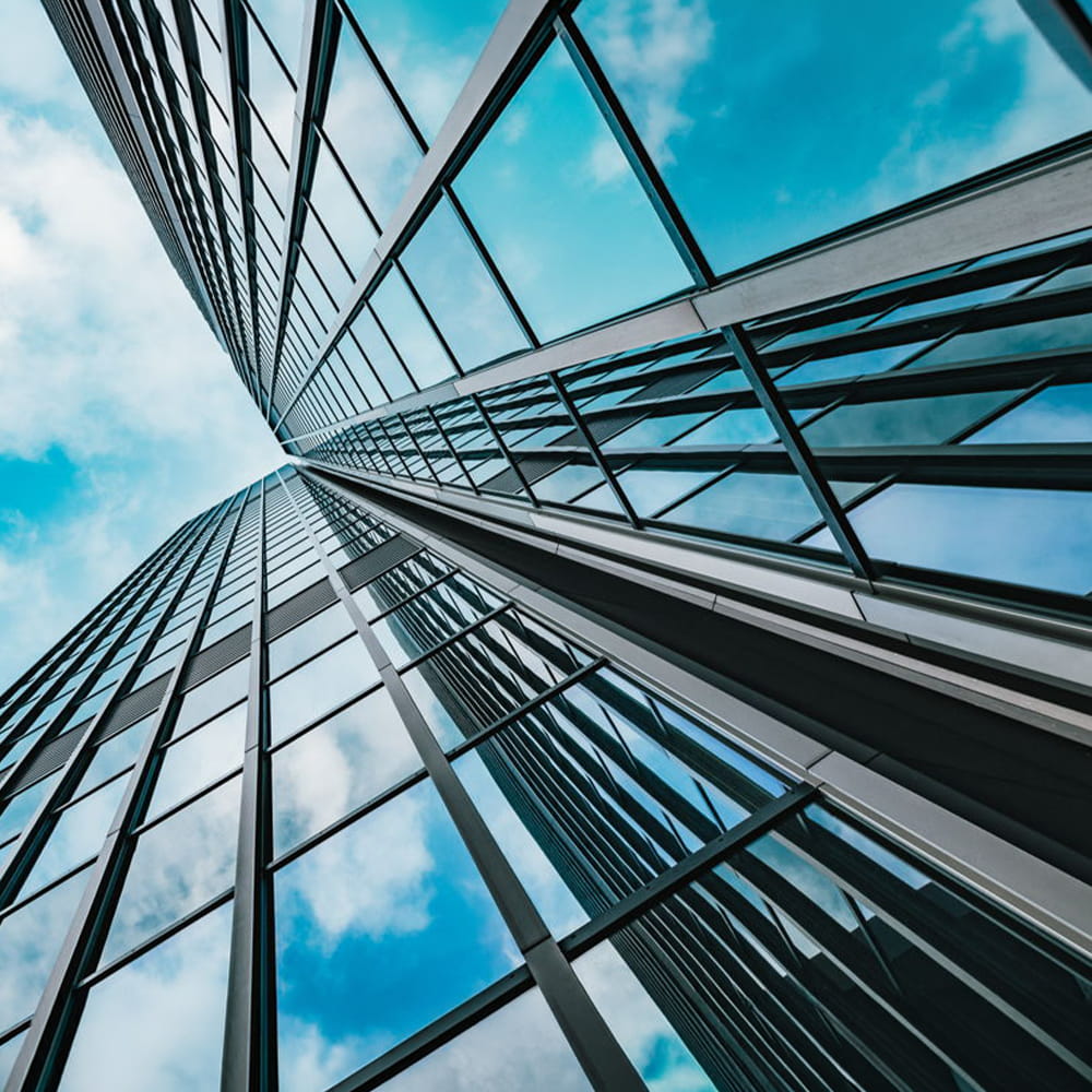 View looking up at a modern glass skyscraper reflecting blue sky and clouds, with sharp lines and geometric patterns converging towards the top of the image.