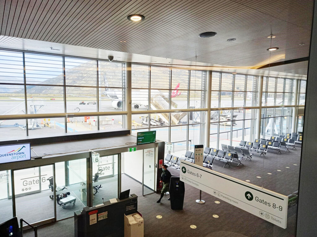 Spacious airport terminal with large windows showing aeroplanes on the tarmac, rows of empty seats, signs for gates 6-9, and mountains visible in the background.