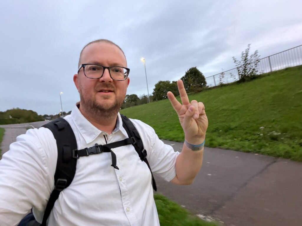 A person wearing glasses, a white shirt, and a black backpack makes a peace sign with their hand while walking on a paved path next to a grassy hill under a cloudy sky.