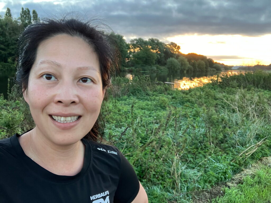 A woman smiles while taking a selfie outdoors with greenery and a river behind her. The sun is setting, casting a warm glow over the landscape and trees in the background.