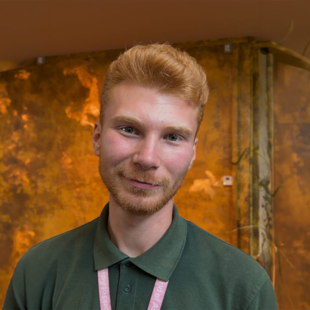 A young man with light skin and reddish-blond hair and beard, wearing a green polo shirt and a lanyard, stands in front of a golden, textured background, smiling gently at the camera.