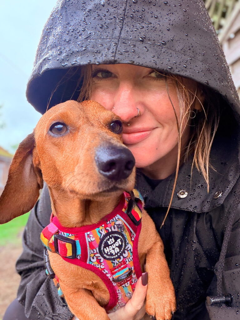 A woman in a rain jacket with a hood smiles while holding a small brown dachshund wearing a colorful harness. Both appear close and happy, with raindrops visible on the womans jacket.