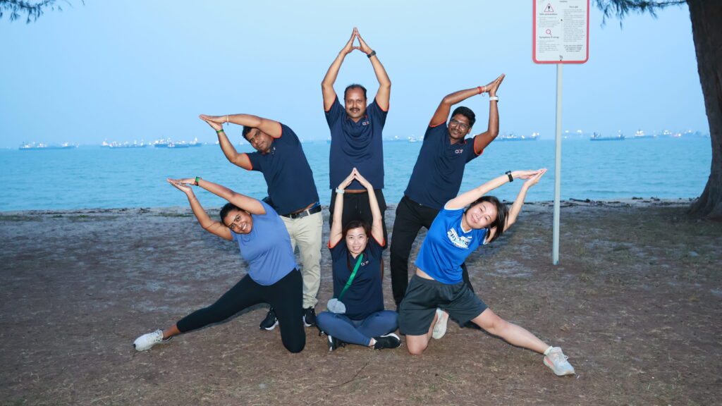 Six people outdoors pose in a playful group, forming a star shape with their arms. The background shows water, a tree, and a sign. They appear happy and coordinated, sitting and kneeling on grass near the shoreline.
