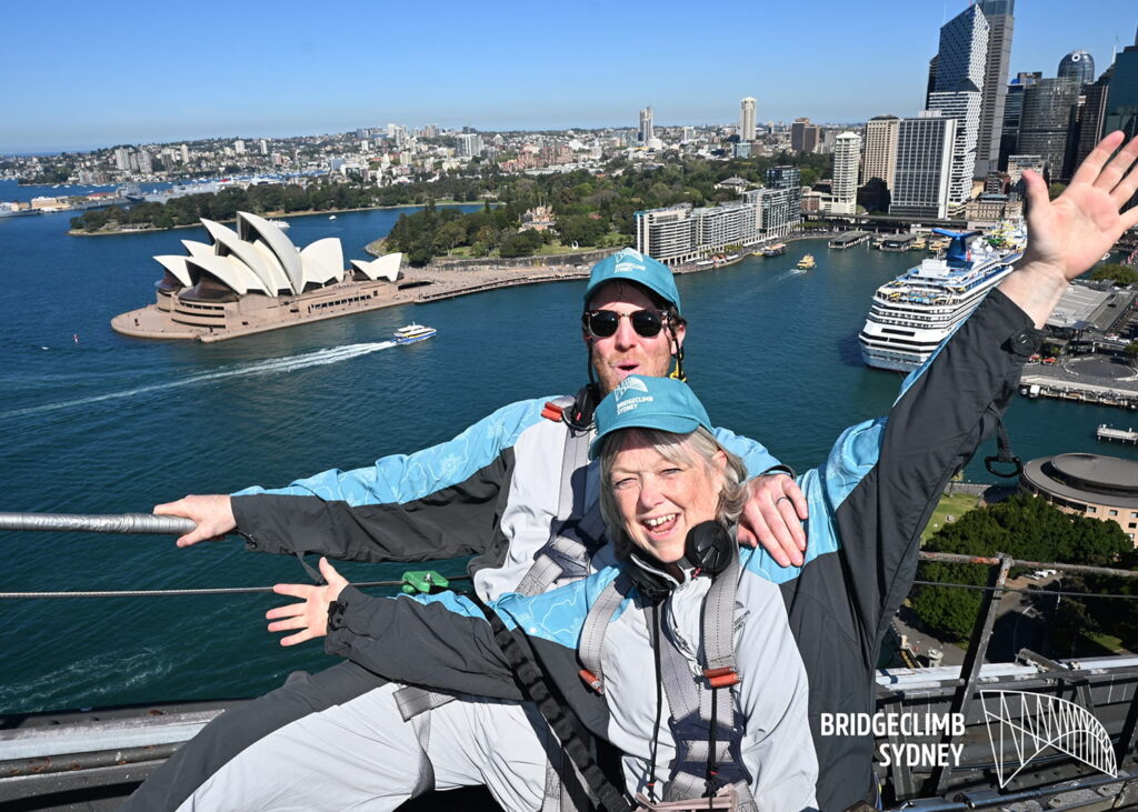 Two people in climbing suits pose joyfully atop Sydney Harbour Bridge, overlooking Sydney Opera House, skyscrapers, and a docked cruise ship. BridgeClimb Sydney logo appears in the lower right corner.