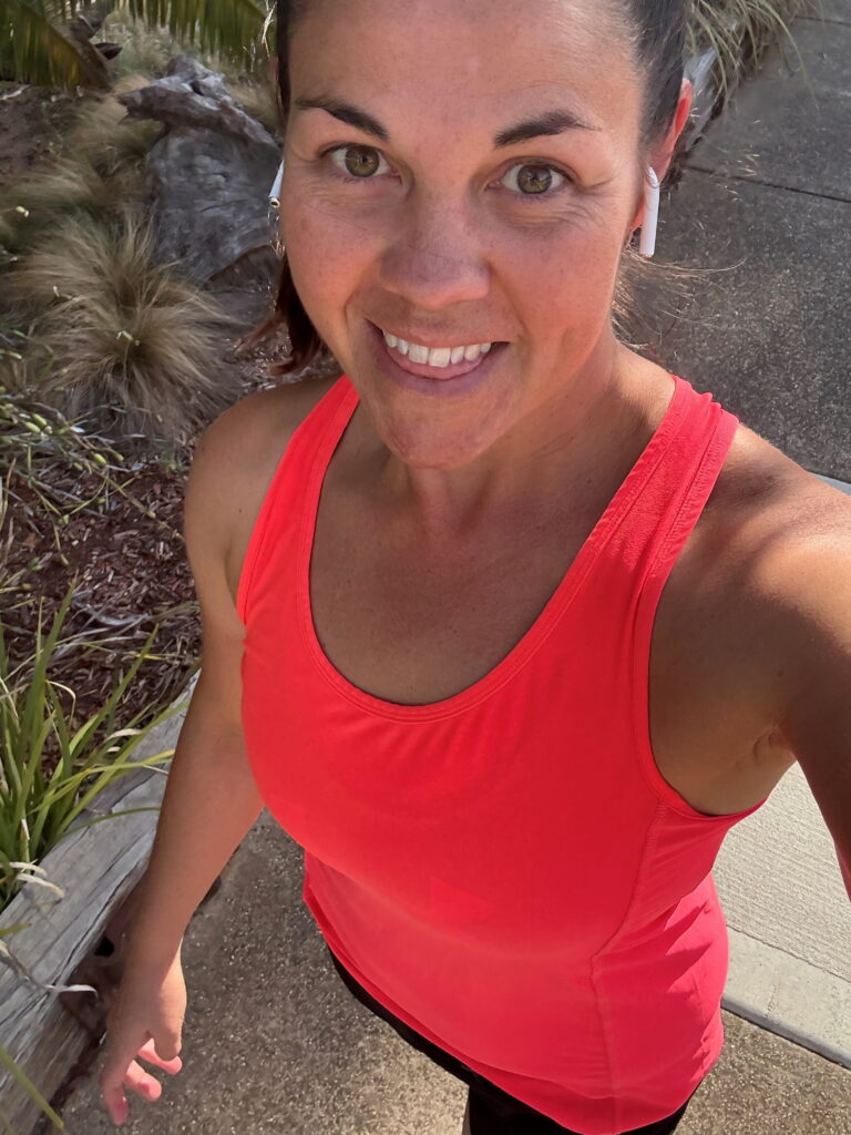 Woman in a bright pink tank top smiles while taking a selfie outdoors, with greenery, pavement, and sunlight in the background.
