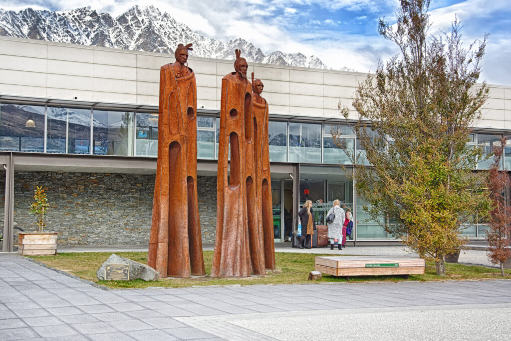 Two tall wooden sculptures of human figures stand on grass outside a modern building, with people near the entrance and snow-capped mountains in the background.