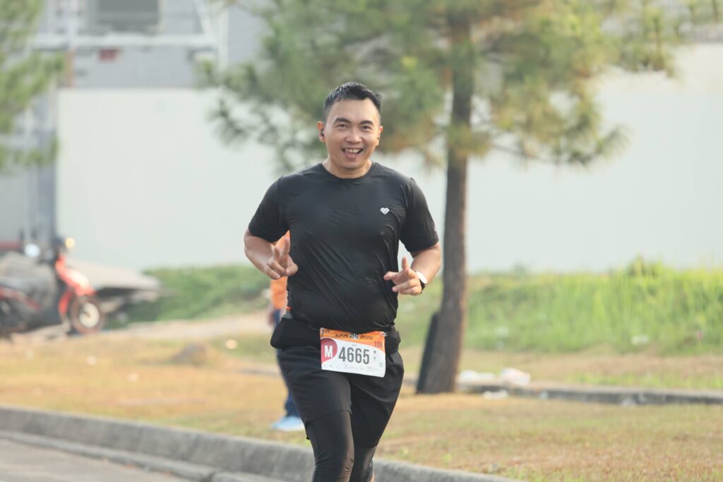 A man wearing black sportswear and a race bib is smiling while running outdoors on a paved path, with trees and grass in the background.