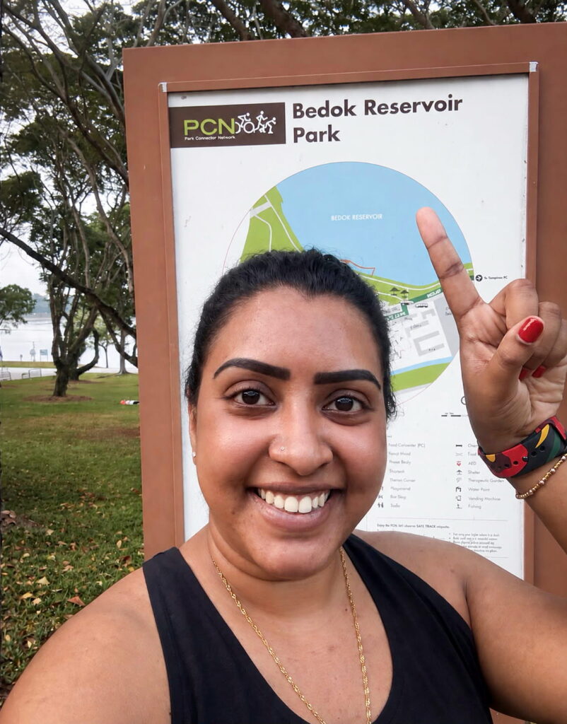 A woman smiles and points upward in front of a Bedok Reservoir Park sign outdoors, with trees and grass in the background.
