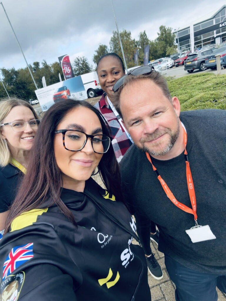 Four people smiling outdoors near cars and a building; one woman in the front holds the camera for a selfie. All wear casual clothes, and one man has an event badge and orange lanyard. Trees and cloudy sky are in the background.