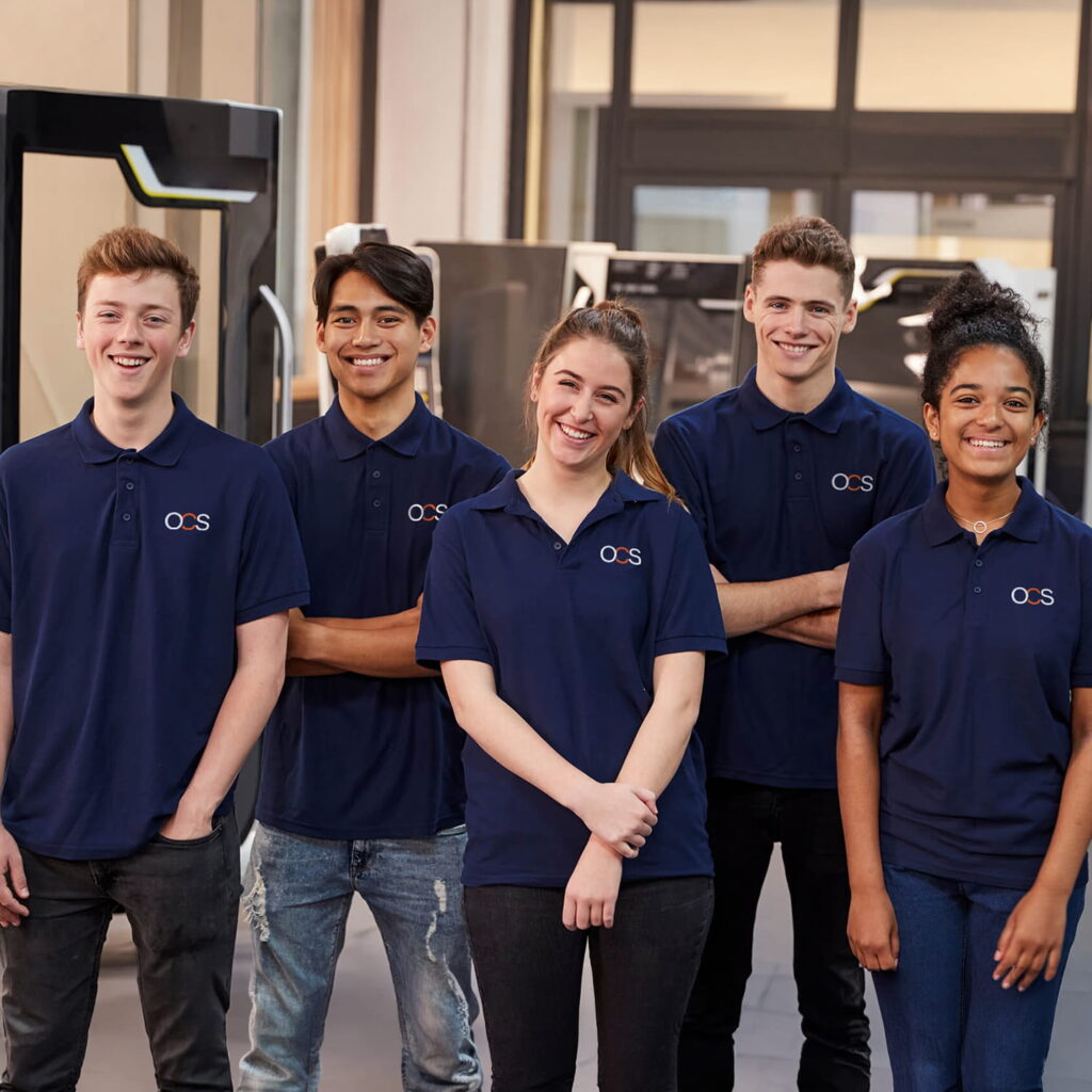Five young adults stand indoors, smiling and wearing matching navy blue polo shirts with a small embroidered logo on the chest. They appear to be part of a team or workplace, posing in a modern office or workspace.