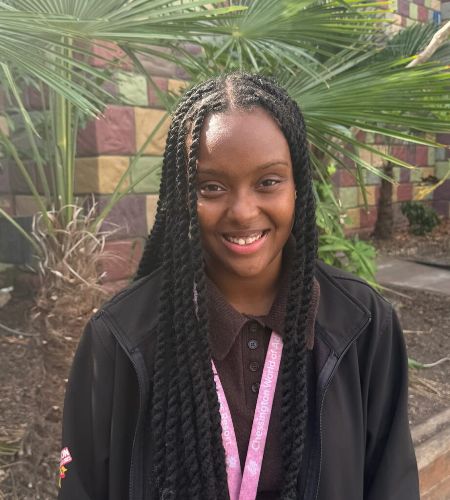 A young woman with long plaits smiles outdoors, wearing a black jacket and a pink lanyard. A colourful tiled wall and green palm leaves are in the background.