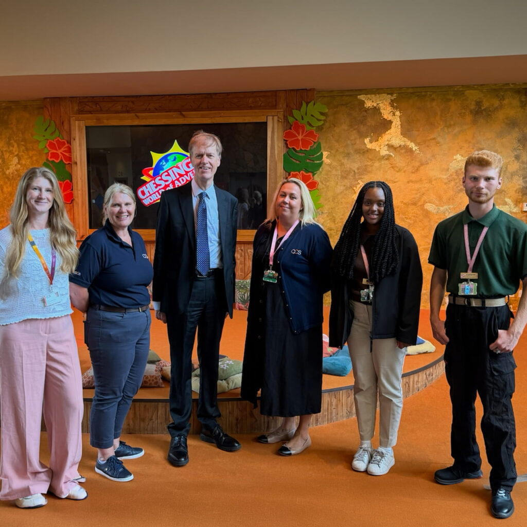Six people stand side by side, smiling at the camera in a colourful indoor setting with wall art and a sign that reads CRESSING WILD KIND. Some wear badges or lanyards.