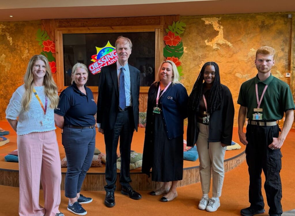 Six people stand indoors on an orange carpet, posing and smiling in front of a TV screen displaying a colourful CBeebies logo. The background features decorative wall art and large red flowers.