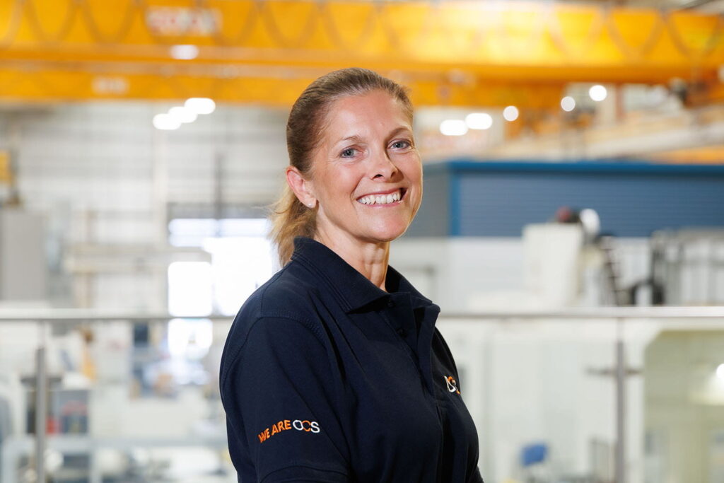 A smiling woman in a navy blue polo shirt stands indoors in an industrial or factory setting, with bright yellow cranes and machinery blurred in the background.