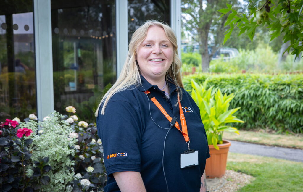 A woman wearing a navy CAREQOS shirt and orange lanyard stands smiling outside near plants and a building with large windows.