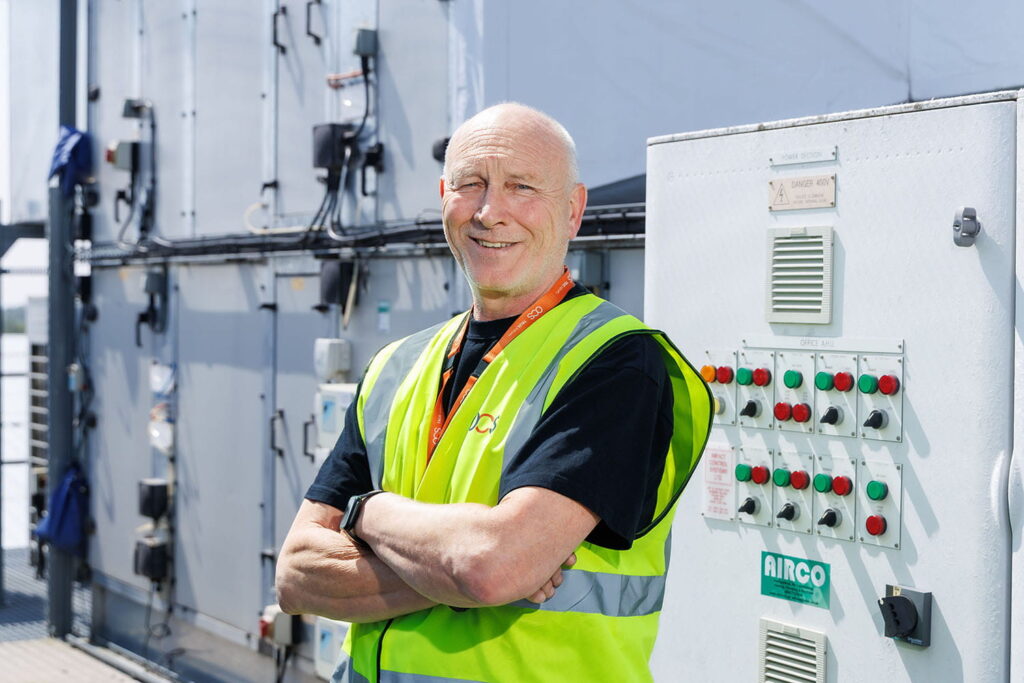 Bald man in a yellow safety vest stands smiling with arms folded in front of industrial equipment and control panels outdoors.