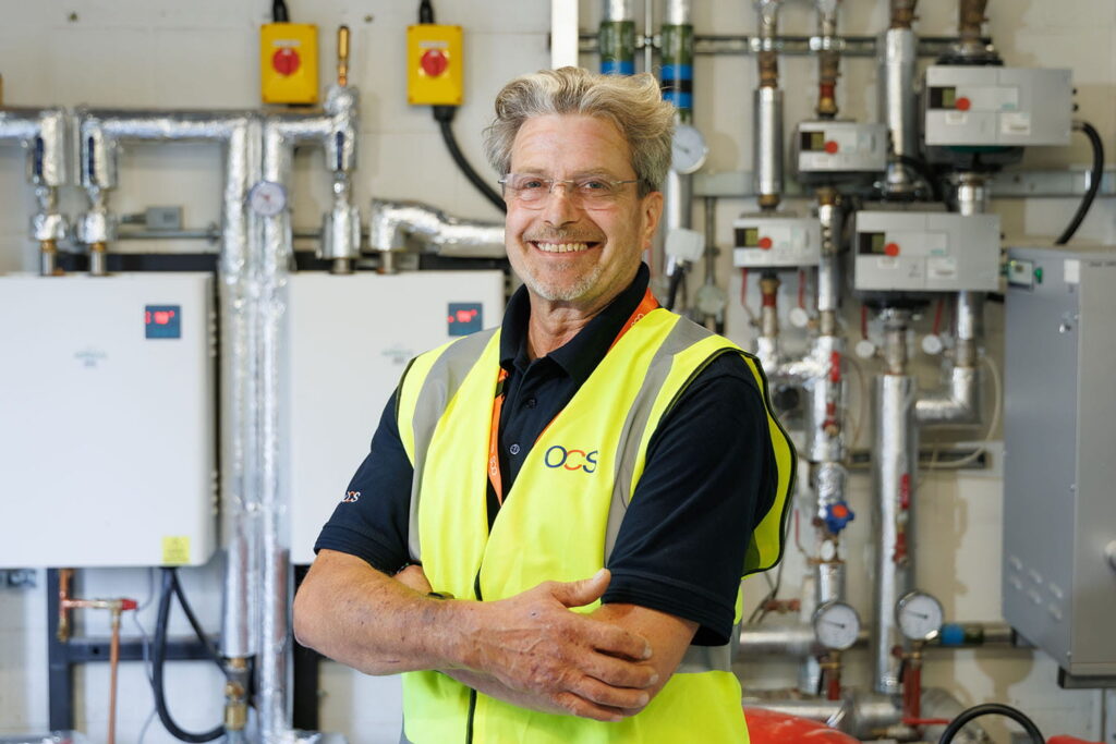 Smiling man with glasses and grey hair wearing a yellow safety vest stands in front of industrial pipes and equipment in a utility room.