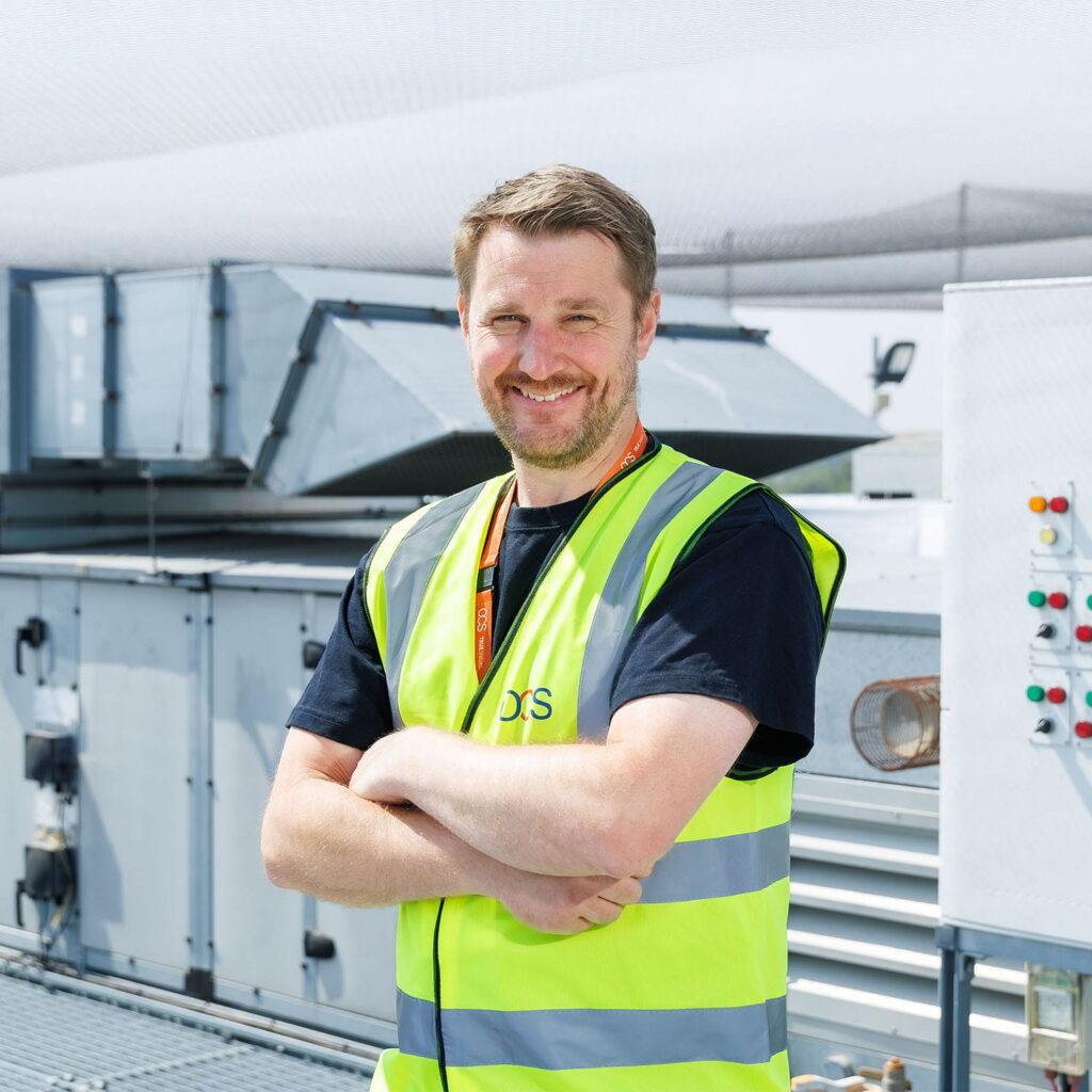 A man in a yellow safety vest stands with arms crossed, smiling on an industrial rooftop near ventilation units and control panels.