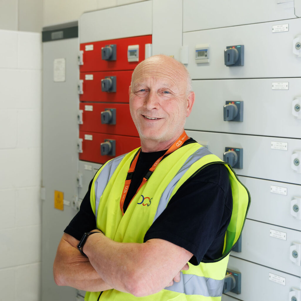 Smiling man in a yellow safety vest stands with arms crossed in front of electrical control panels, wearing a black shirt and orange lanyard.