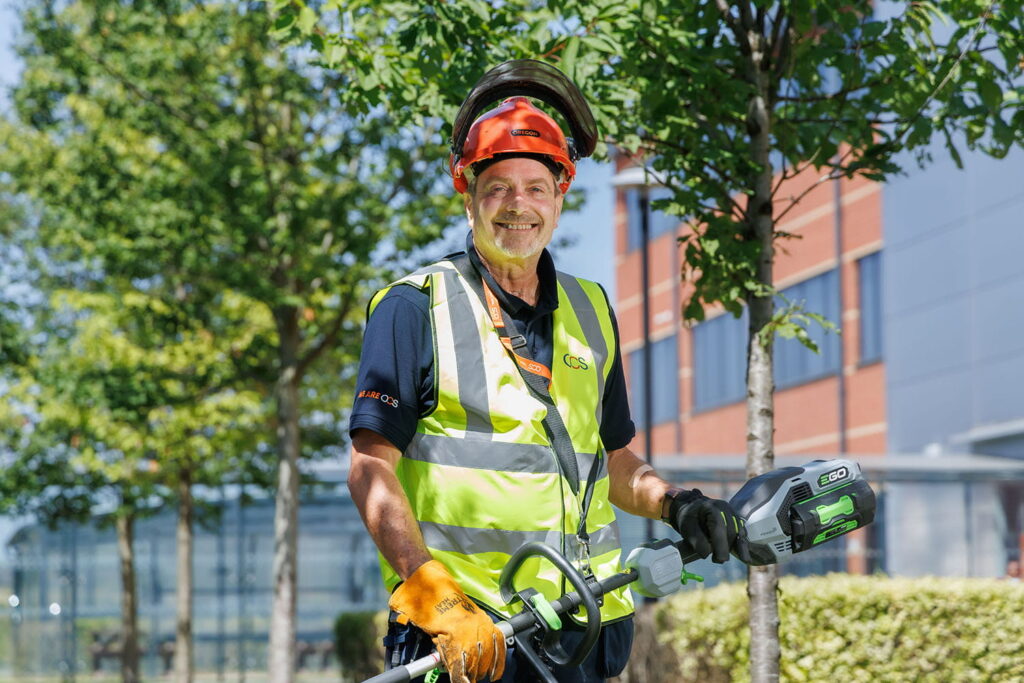 A smiling worker in a safety vest, helmet, and gloves holds a power trimmer outdoors near trees and a modern building on a sunny day.