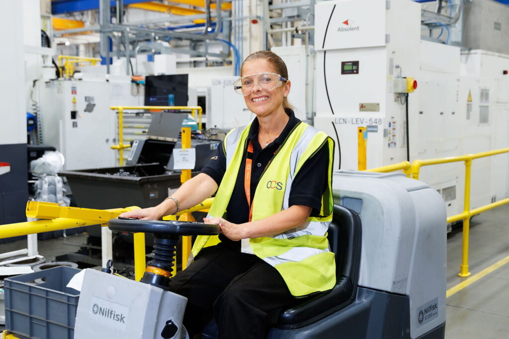A woman wearing safety glasses and a high-visibility vest smiles whilst operating a floor cleaning machine in an industrial factory setting.