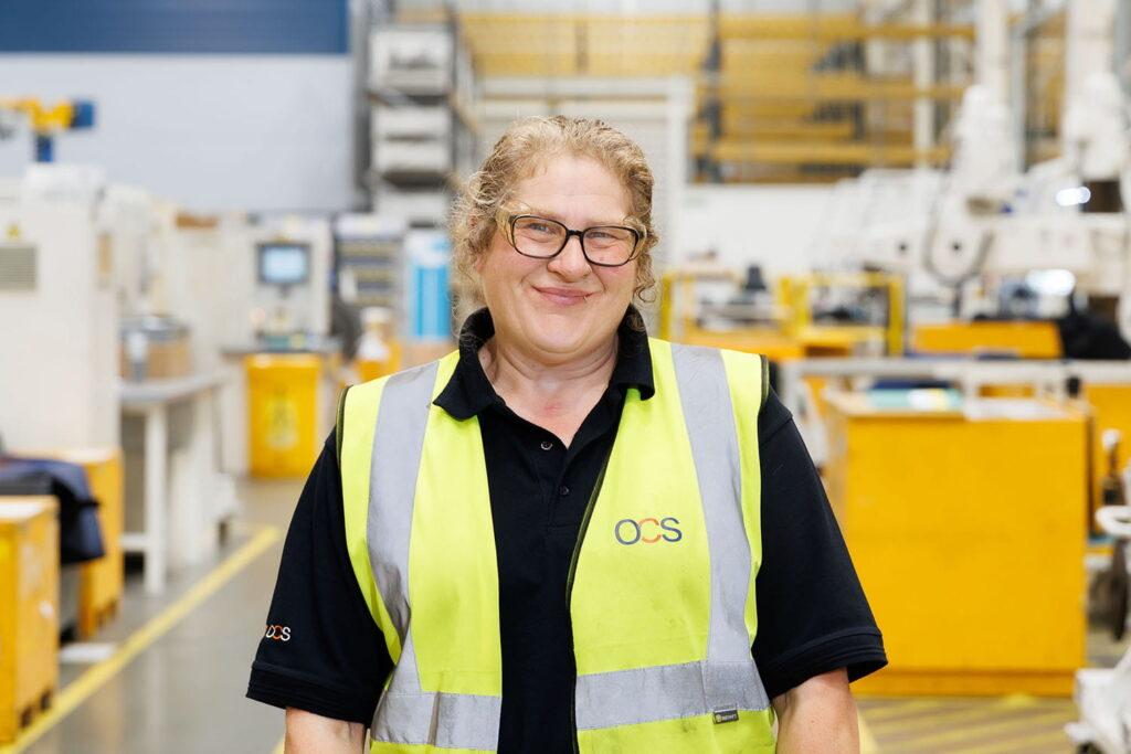 A smiling woman in glasses and a yellow safety vest stands in a brightly lit industrial warehouse with yellow workstations and machinery in the background.