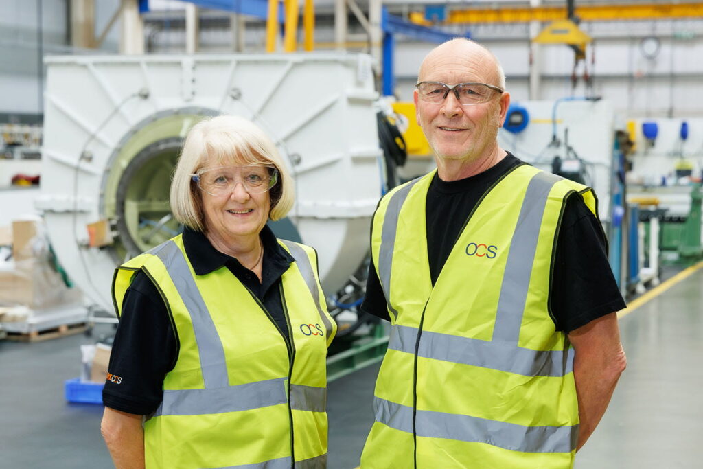 Two older adults wearing glasses, black shirts, and yellow high-visibility vests stand smiling in a bright industrial facility with machinery and equipment in the background.