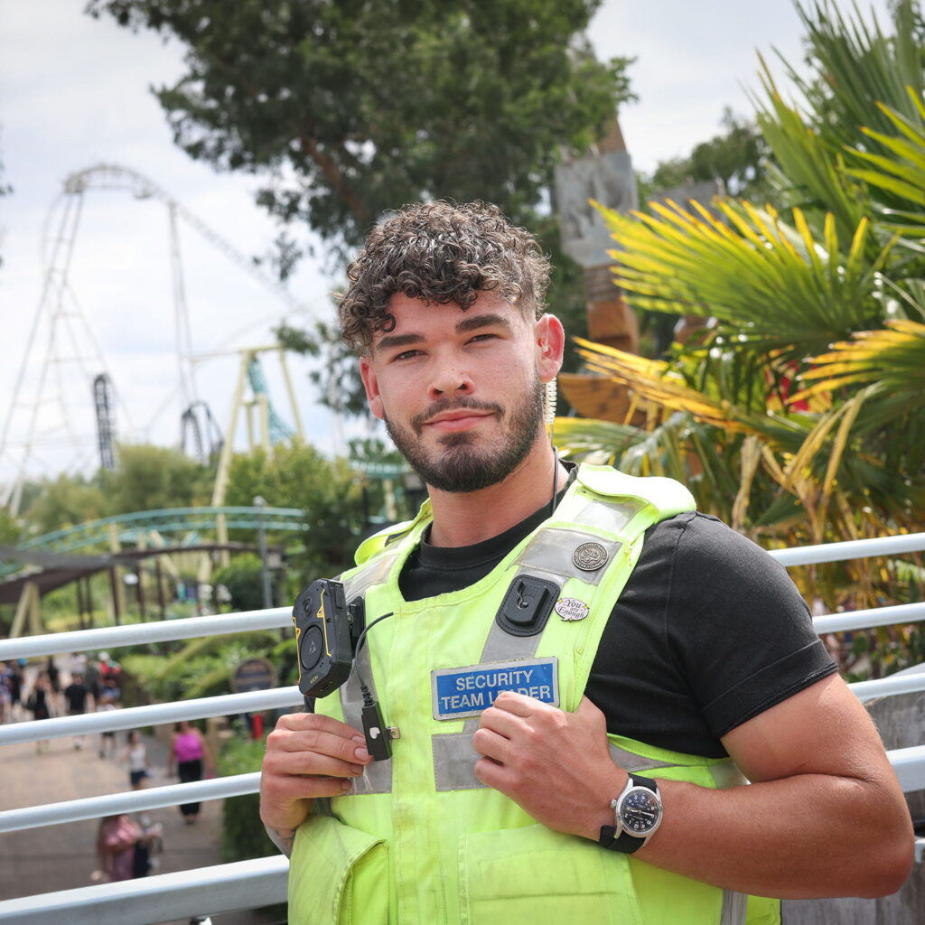 A security team leader wearing a high-visibility vest stands outdoors near metal railings, with a rollercoaster and palm trees in the background.