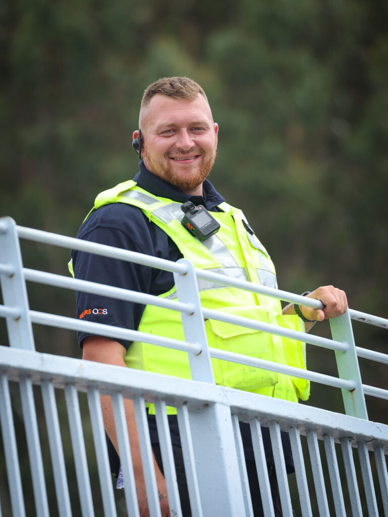 A smiling man wearing a high-visibility safety vest and radio earpiece stands outdoors by a metal railing, with greenery blurred in the background.