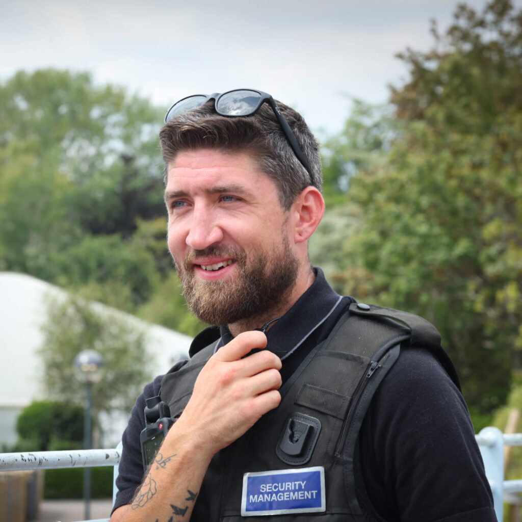 A bearded man in a black Security Management vest holds a radio and smiles outdoors. Trees and greenery are blurred in the background. Sunglasses rest on his head.