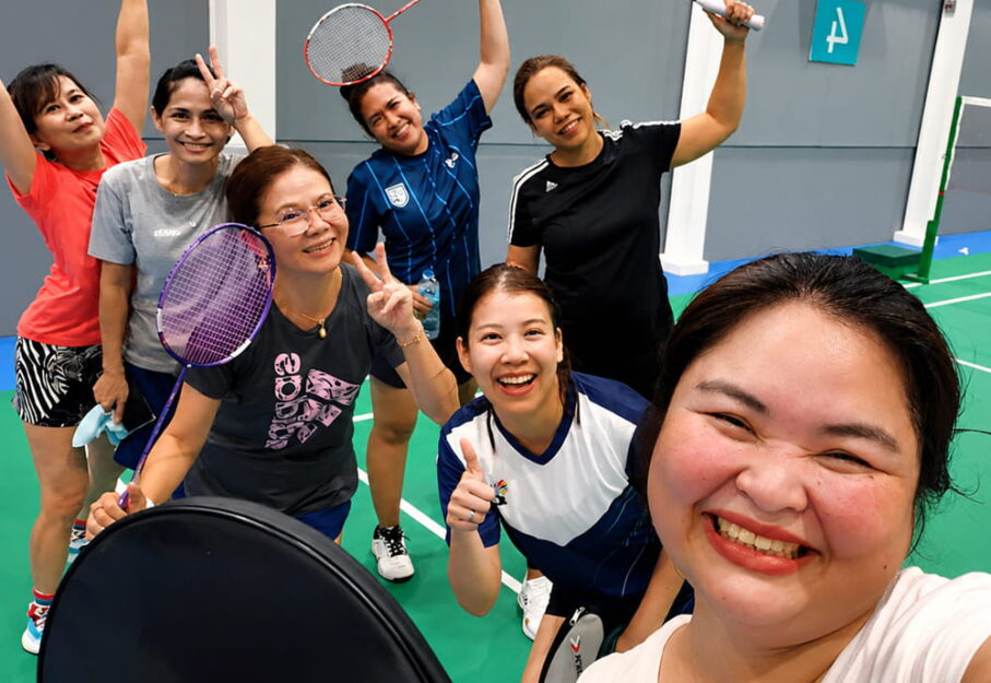 Seven women pose and smile on an indoor badminton court, holding rackets and making peace signs, appearing happy and energetic after playing a game together.