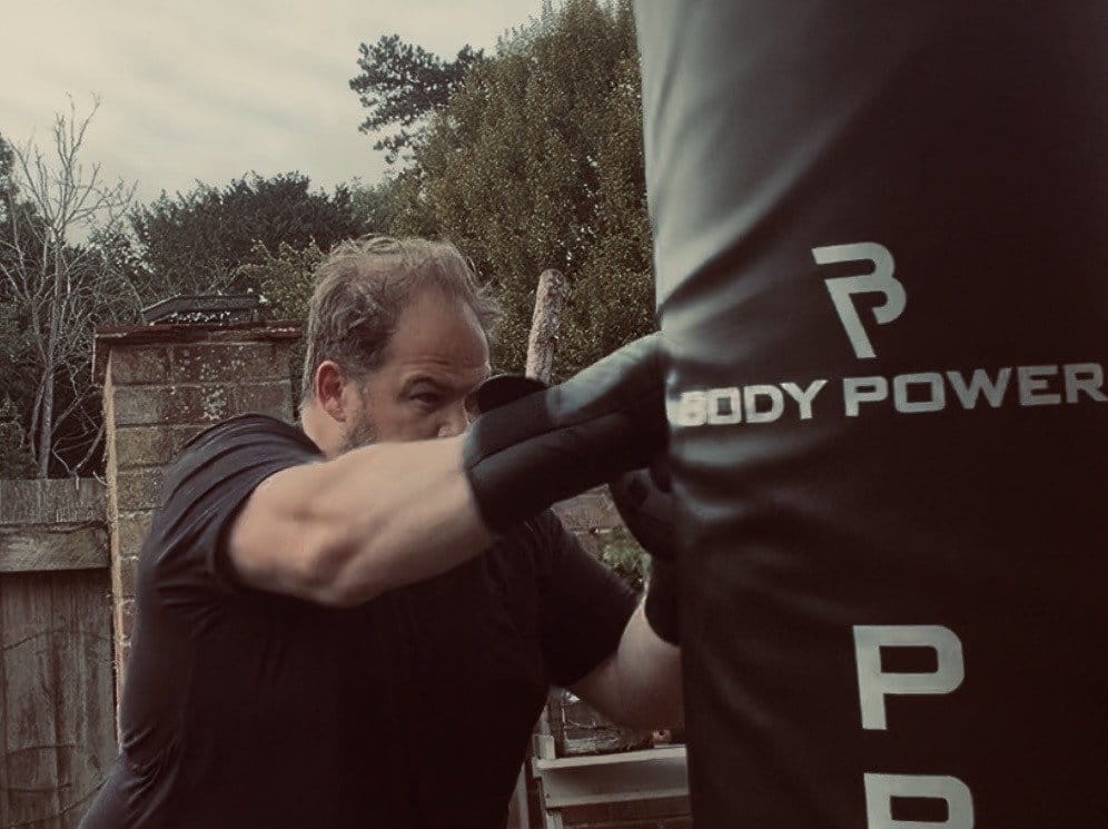 A person wearing black boxing gloves punches a Body Power punching bag outdoors, with trees and a wooden fence in the background.