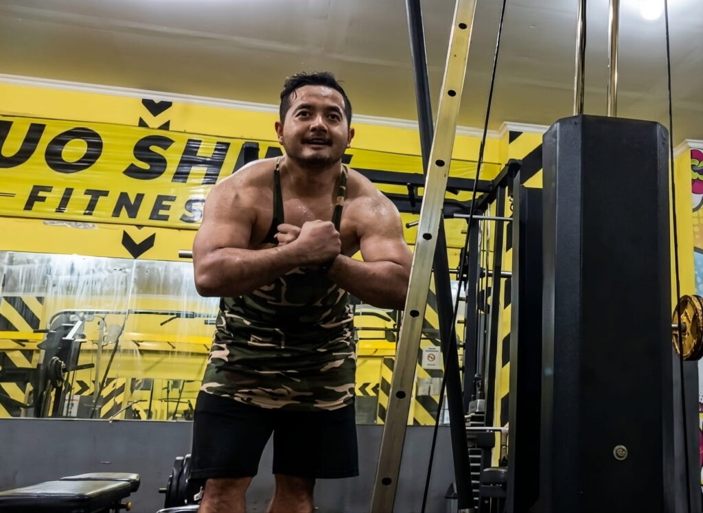 A man in a camouflage tank top flexes his muscles and smiles inside a brightly lit gym with yellow and black walls. Fitness equipment is visible in the background.
