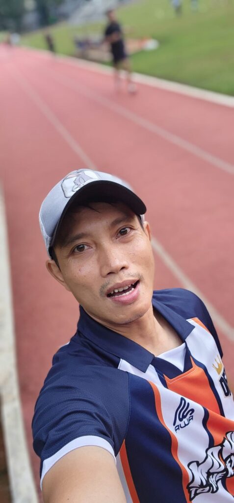 A man wearing a cap and a sports jersey takes a selfie on a red running track. The background shows a grassy area and another person walking on the track.
