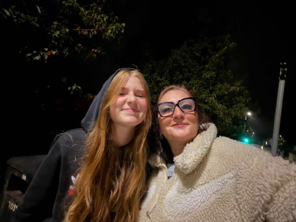 Two people stand close together outside at night, smiling at the camera. One has long red hair and wears a hoodie, the other has glasses and wears a fuzzy jacket. There are trees and streetlights in the background.
